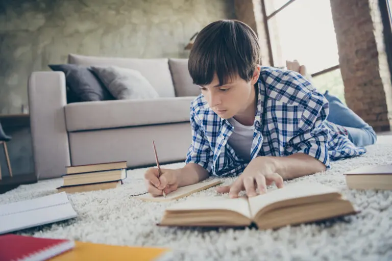 A young boy lies on a rug, writing in a notebook, surrounded by open books and notebooks, in a cozy, bright living room.