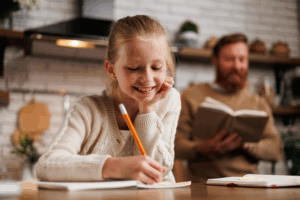 Smiling girl writing in a notebook at a kitchen table, with an adult reading in the background.