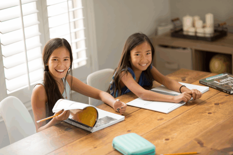 Two smiling girls sitting at a wooden table, working in spiral-bound science notebooks at home.
